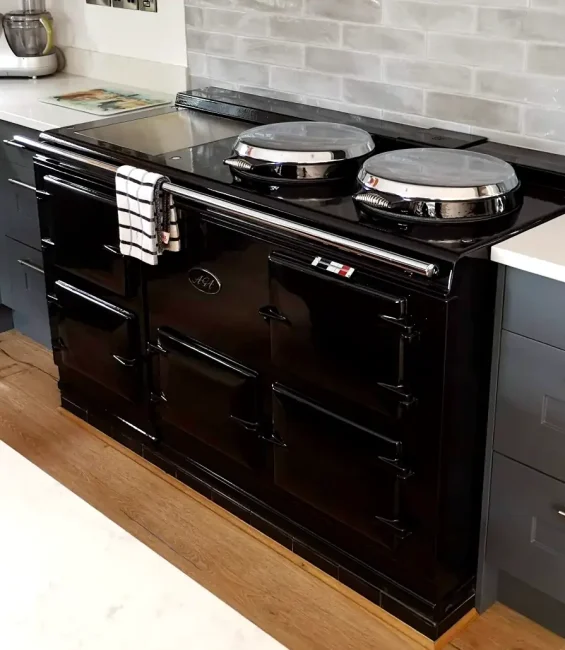 Glossy black aga cooker in a sleek modern kitchen with grey cabinetry and stainless steel cookware. Displayed by John Wray Range Cookers.