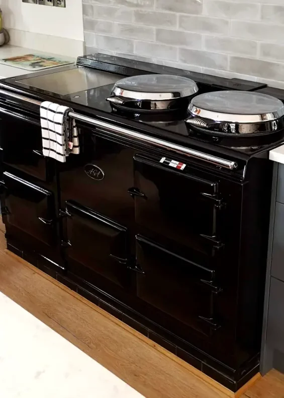 Glossy black aga cooker in a sleek modern kitchen with grey cabinetry and stainless steel cookware. Displayed by John Wray Range Cookers.